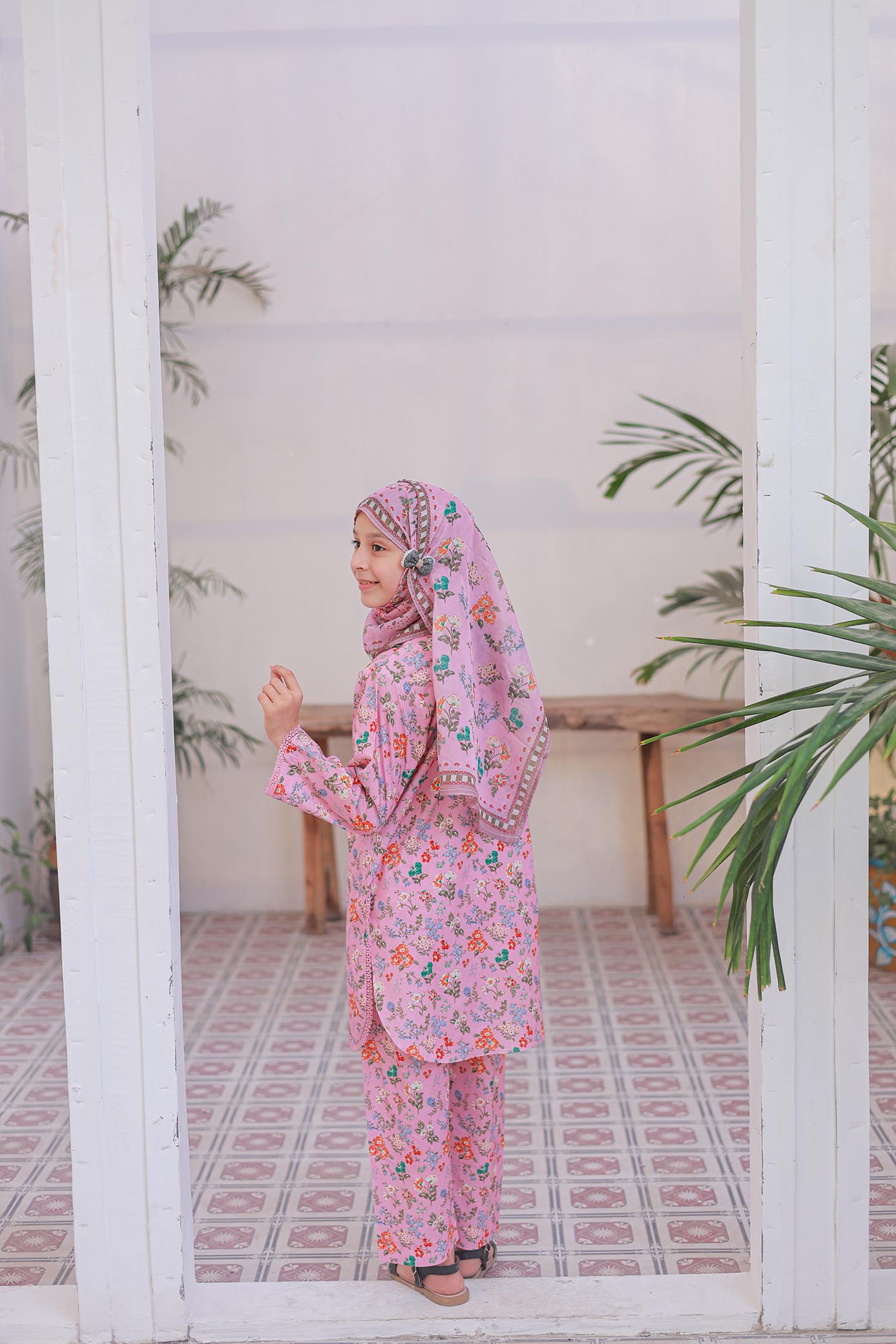Woman in a pink floral outfit standing in a room with plants and a white wall.
