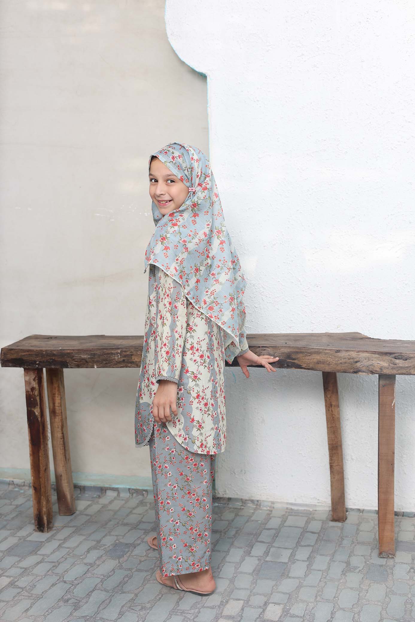 Woman wearing a floral outfit standing behind a wooden bench against a white wall.