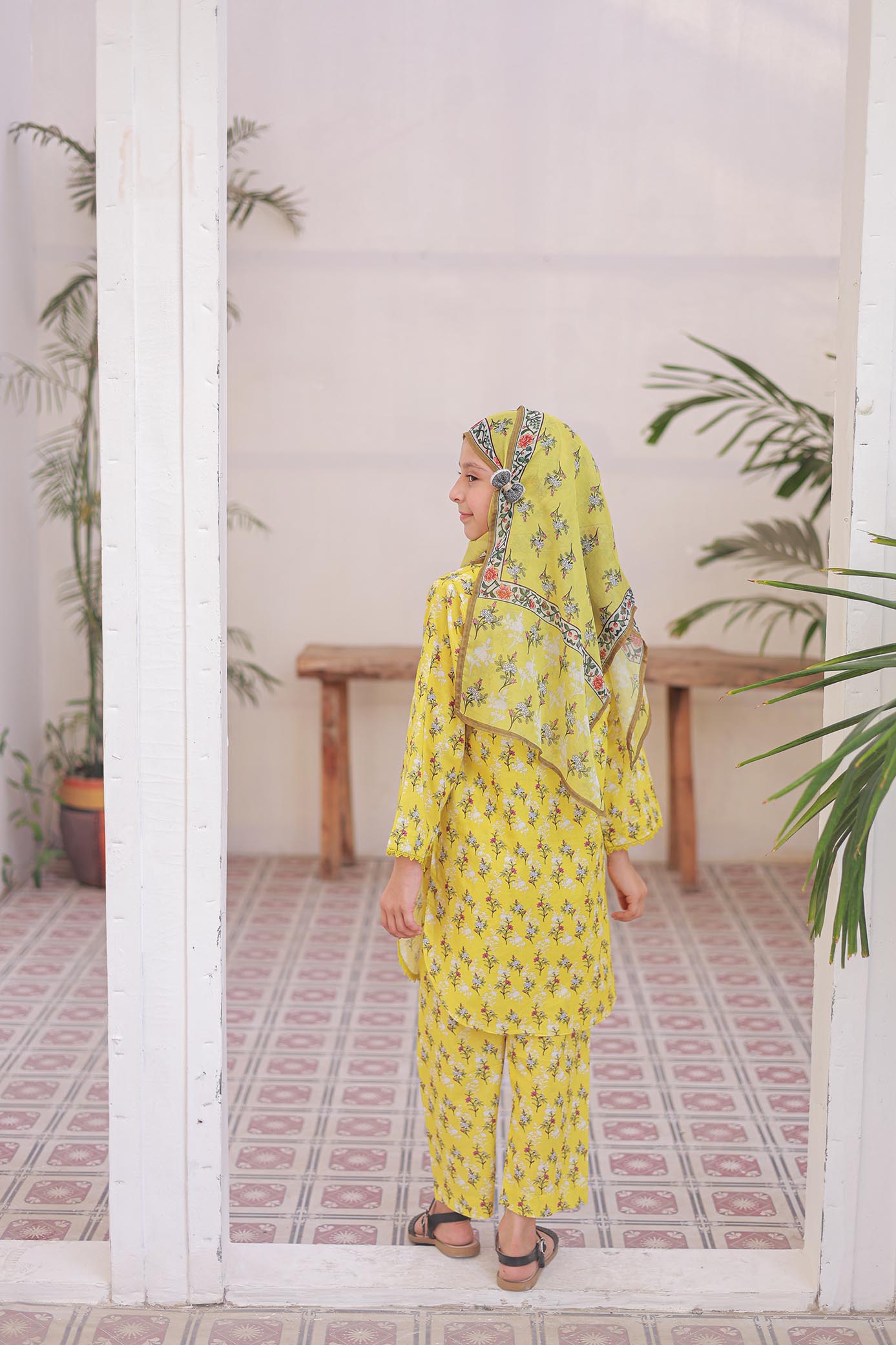 Woman in a yellow traditional outfit standing in a room with plants and a tiled floor.