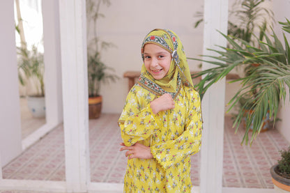 Young girl in a yellow traditional outfit with a patterned headscarf standing in a garden.