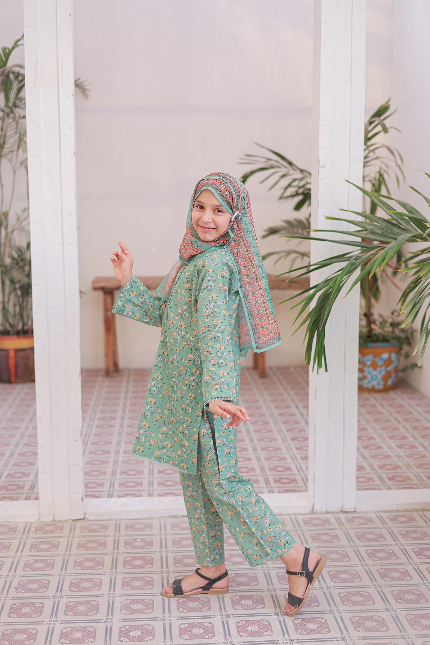 Young girl in a green floral outfit with a headscarf standing in a room with plants.