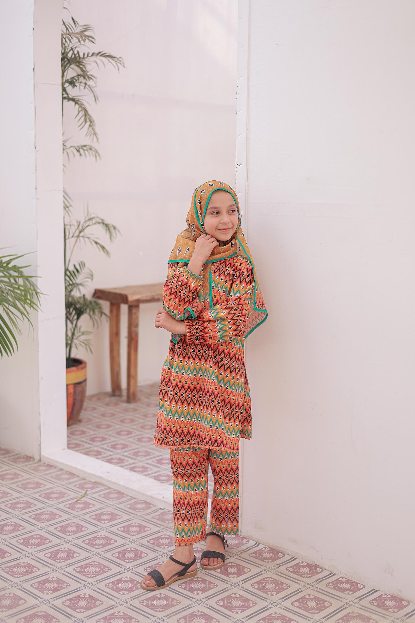 Woman wearing a colorful patterned outfit and hijab indoors with plants and a mirror in the background.