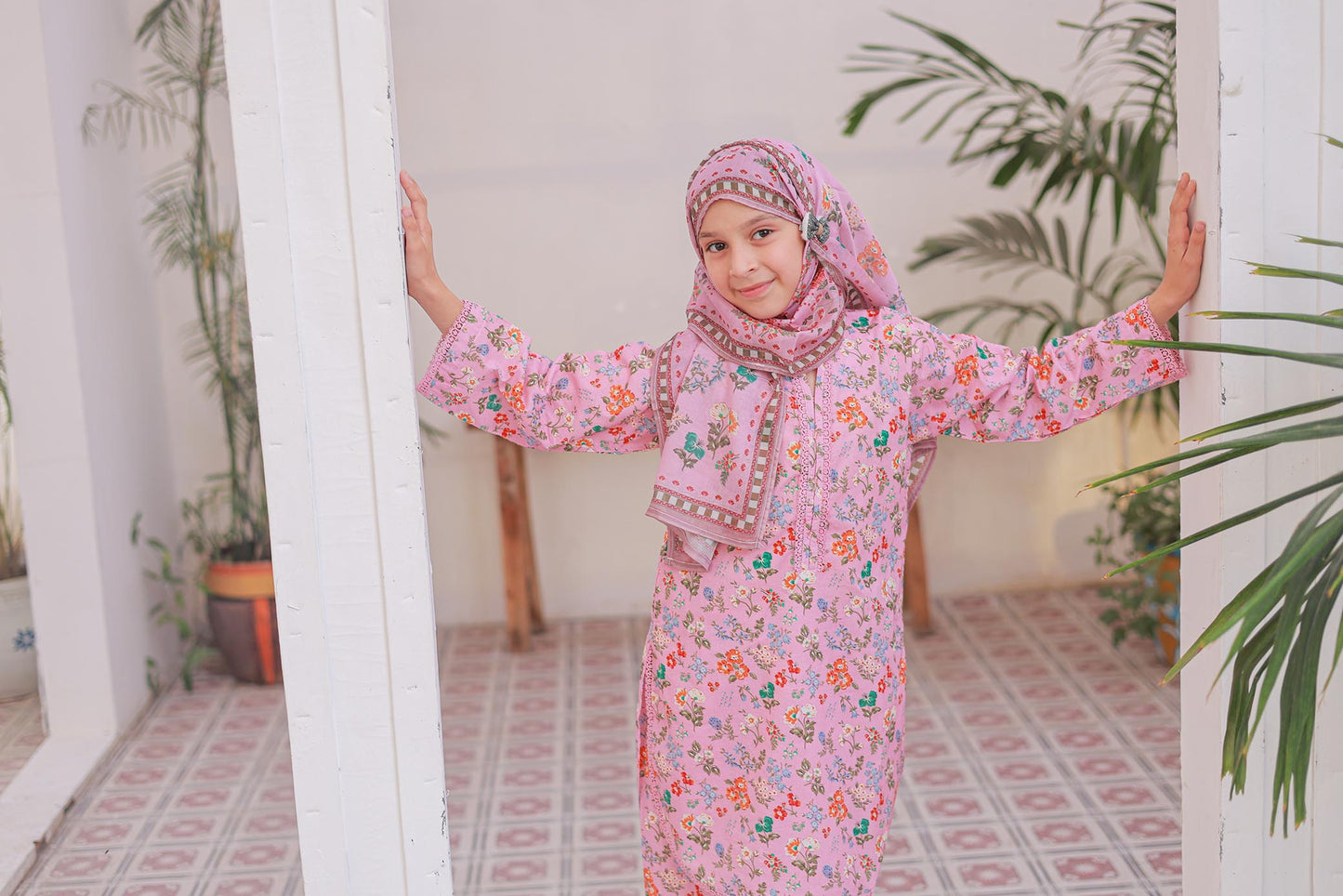 Child in a pink floral dress standing in a room with plants.