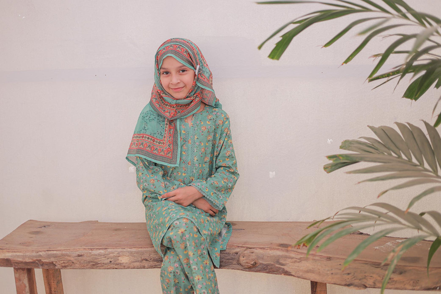 Woman in a green traditional outfit sitting on a wooden bench with plants in the background