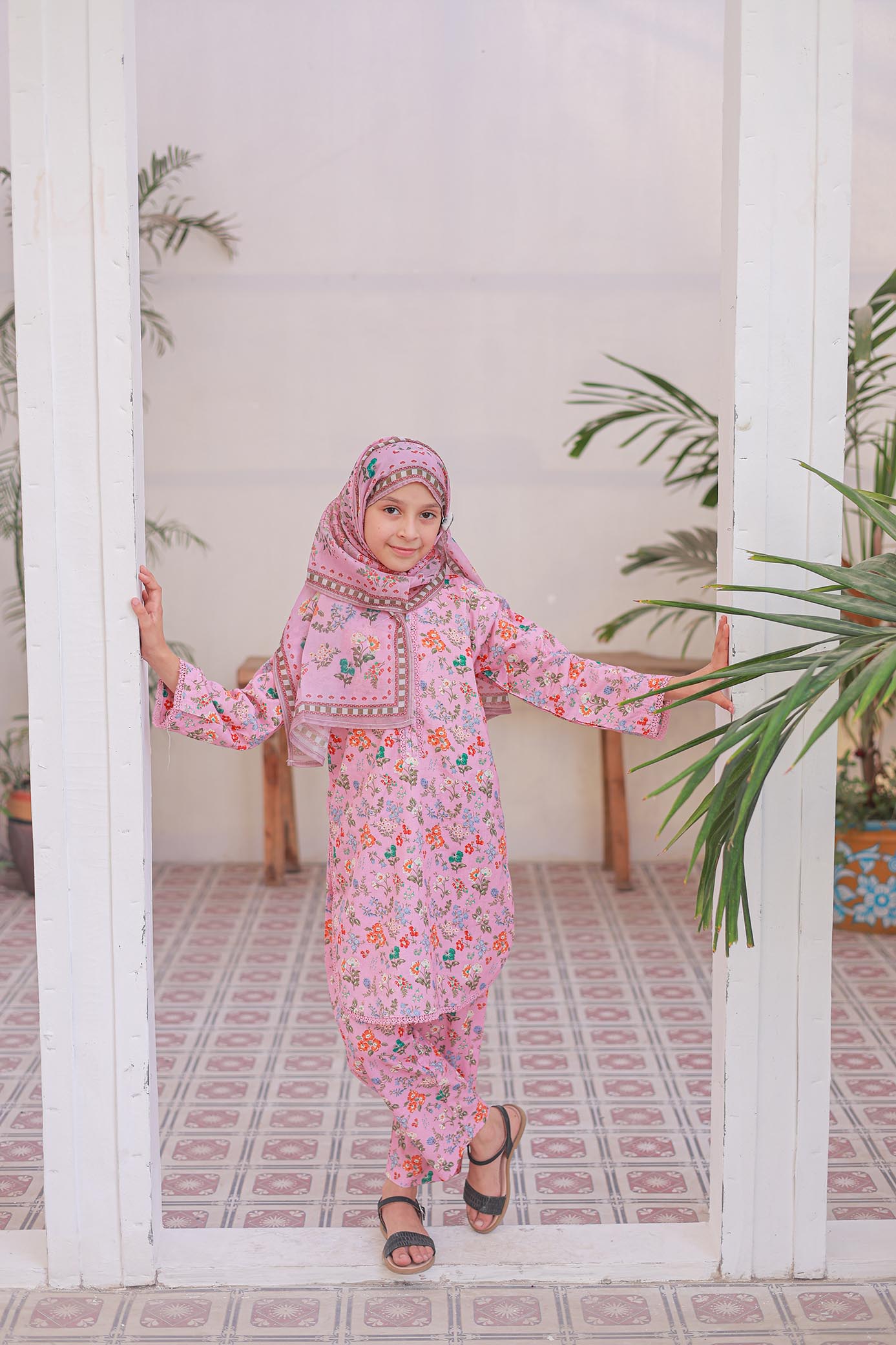 Child in a pink floral outfit standing in a room with plants and a tiled floor.
