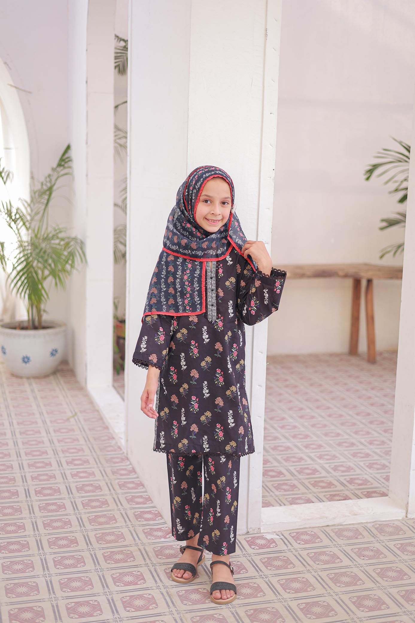Child wearing a dark floral outfit with a matching hijab in a room with plants and a table.