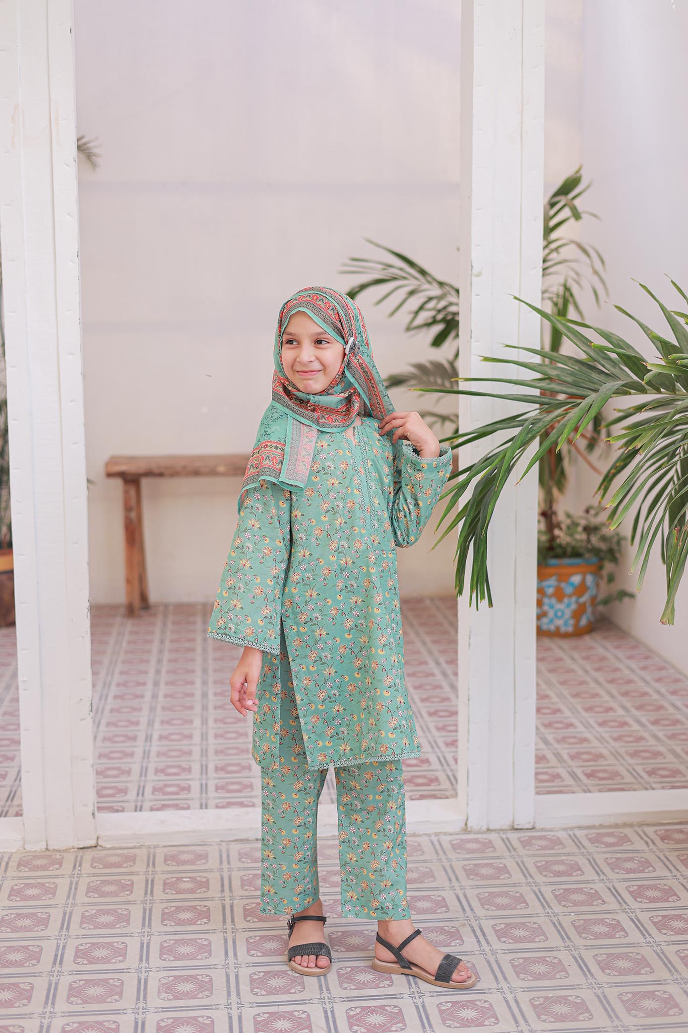 Child wearing a green traditional outfit with a headscarf in a room with white columns and plants.