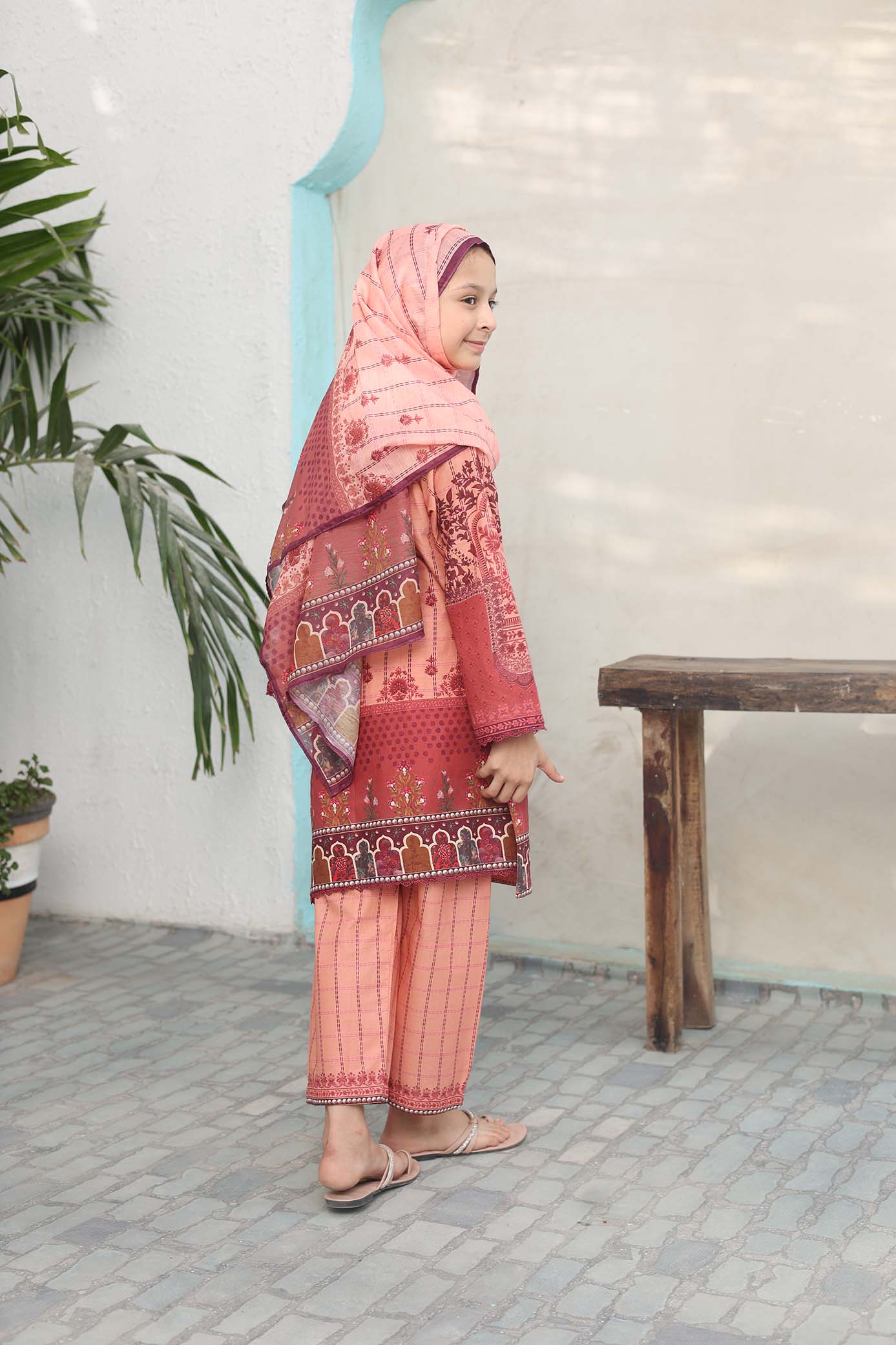 Woman wearing a traditional outfit with a pink hijab and red and pink patterned dress in an indoor setting.