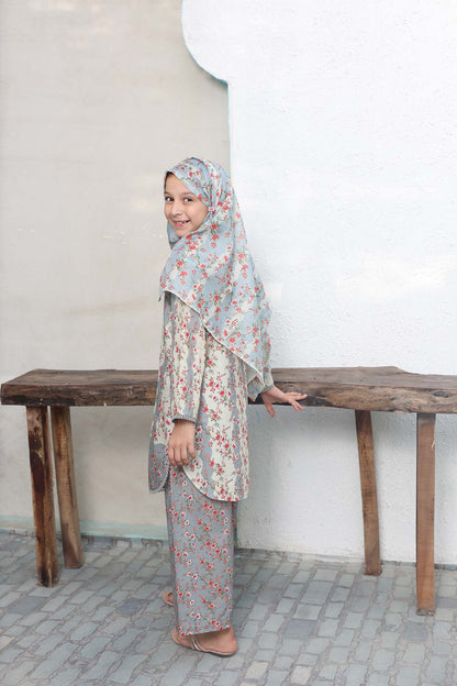 Woman wearing a floral outfit standing behind a wooden bench against a white wall.