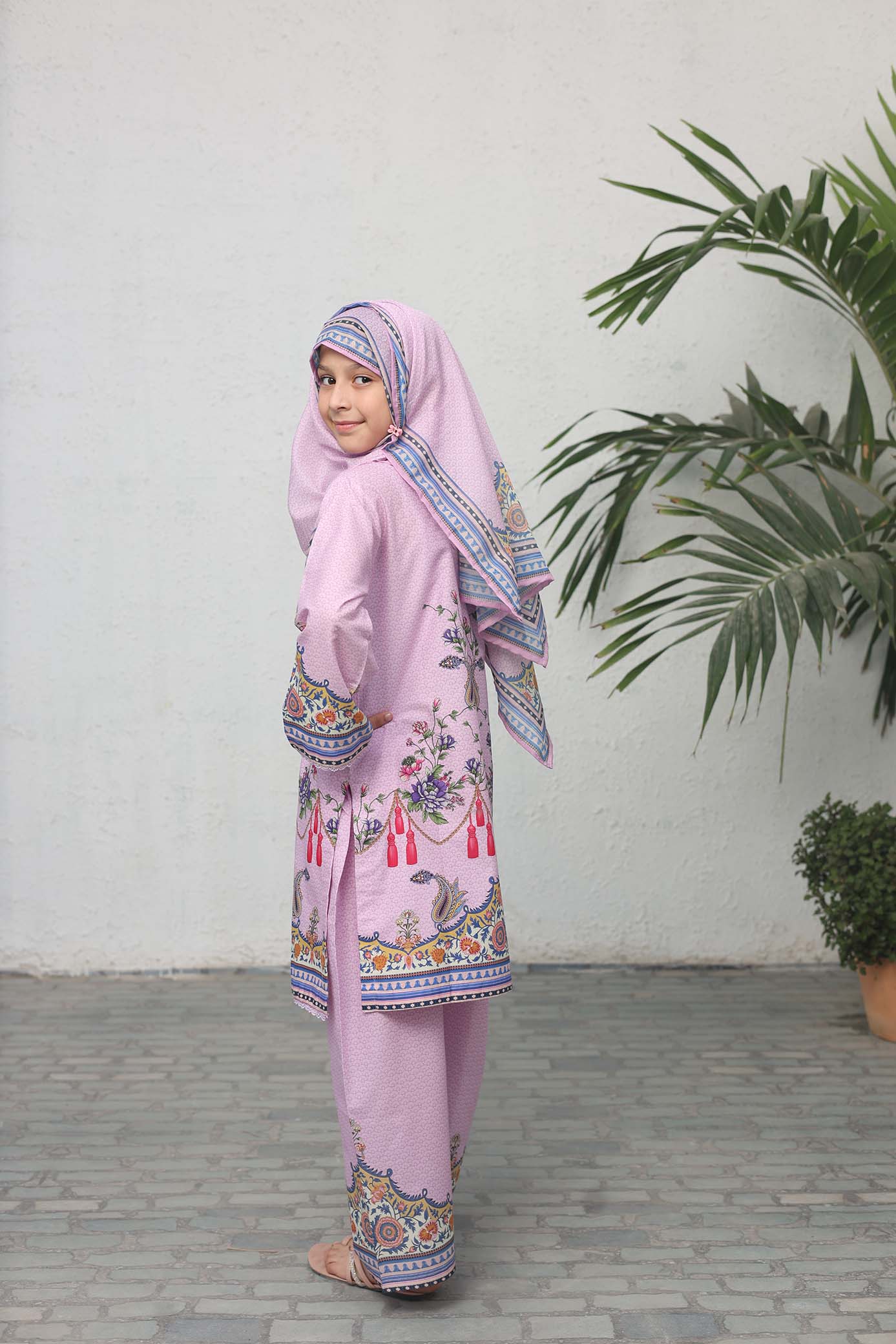 Child wearing a pink traditional outfit with a matching headscarf, standing against a white wall with a plant.