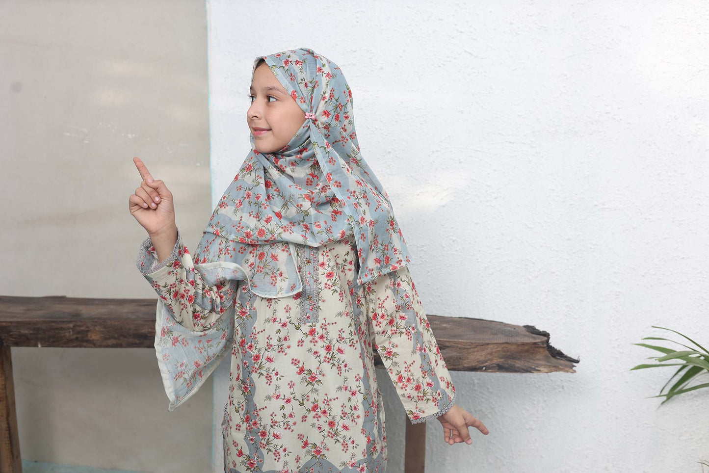 Child wearing a floral dress and headscarf pointing upwards against a white wall.