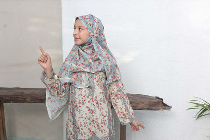 Child wearing a floral dress and headscarf pointing upwards against a white wall.