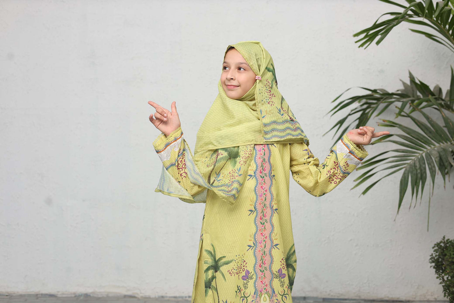 Young girl in a green traditional outfit with a hijab, pointing to the side against a white wall with a plant.