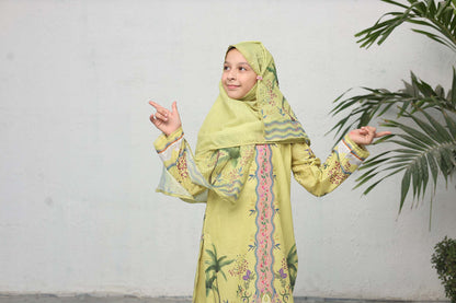 Young girl in a green traditional outfit with a hijab, pointing to the side against a white wall with a plant.