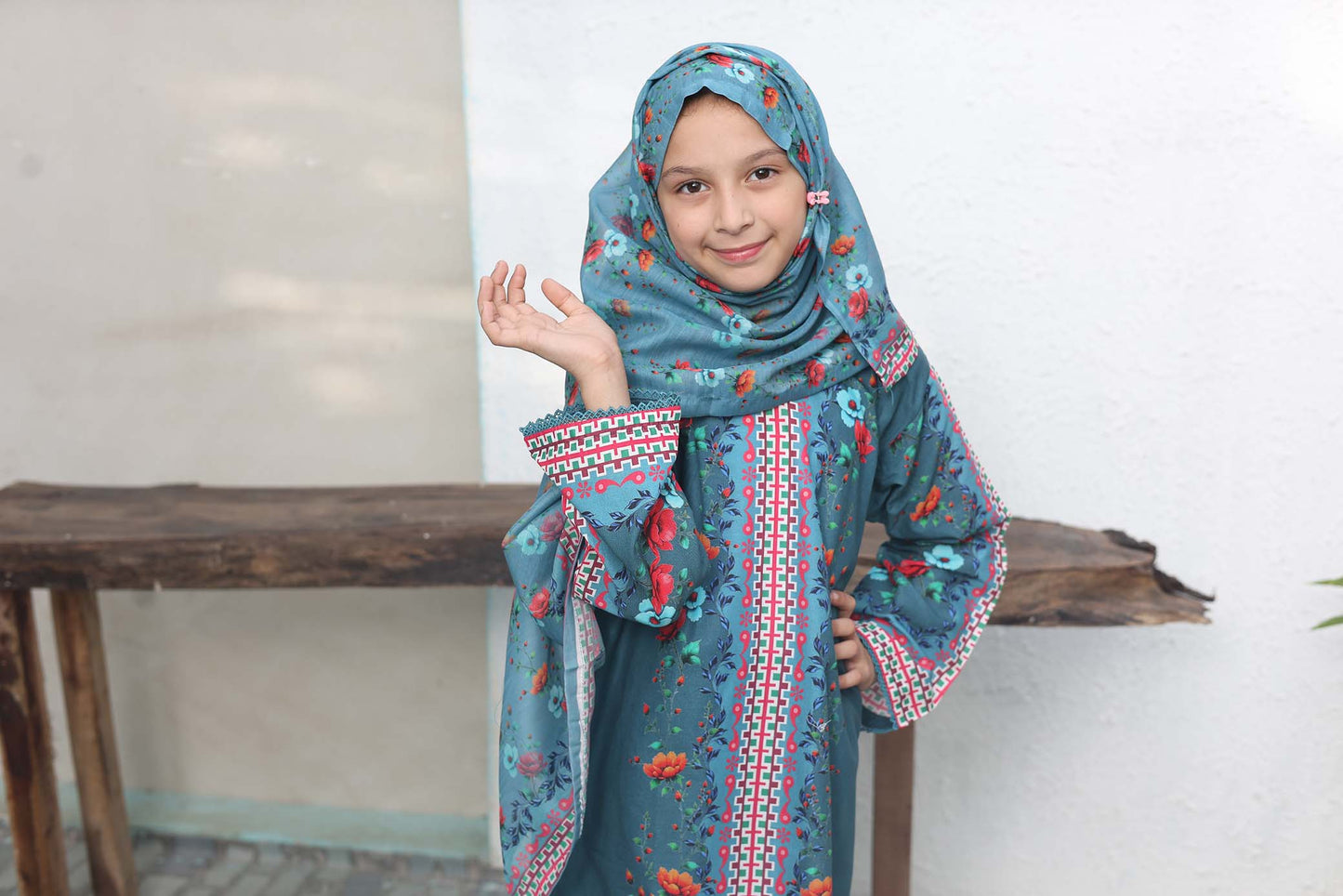 Young girl wearing a blue traditional outfit with a matching hijab, standing in front of a wooden table.