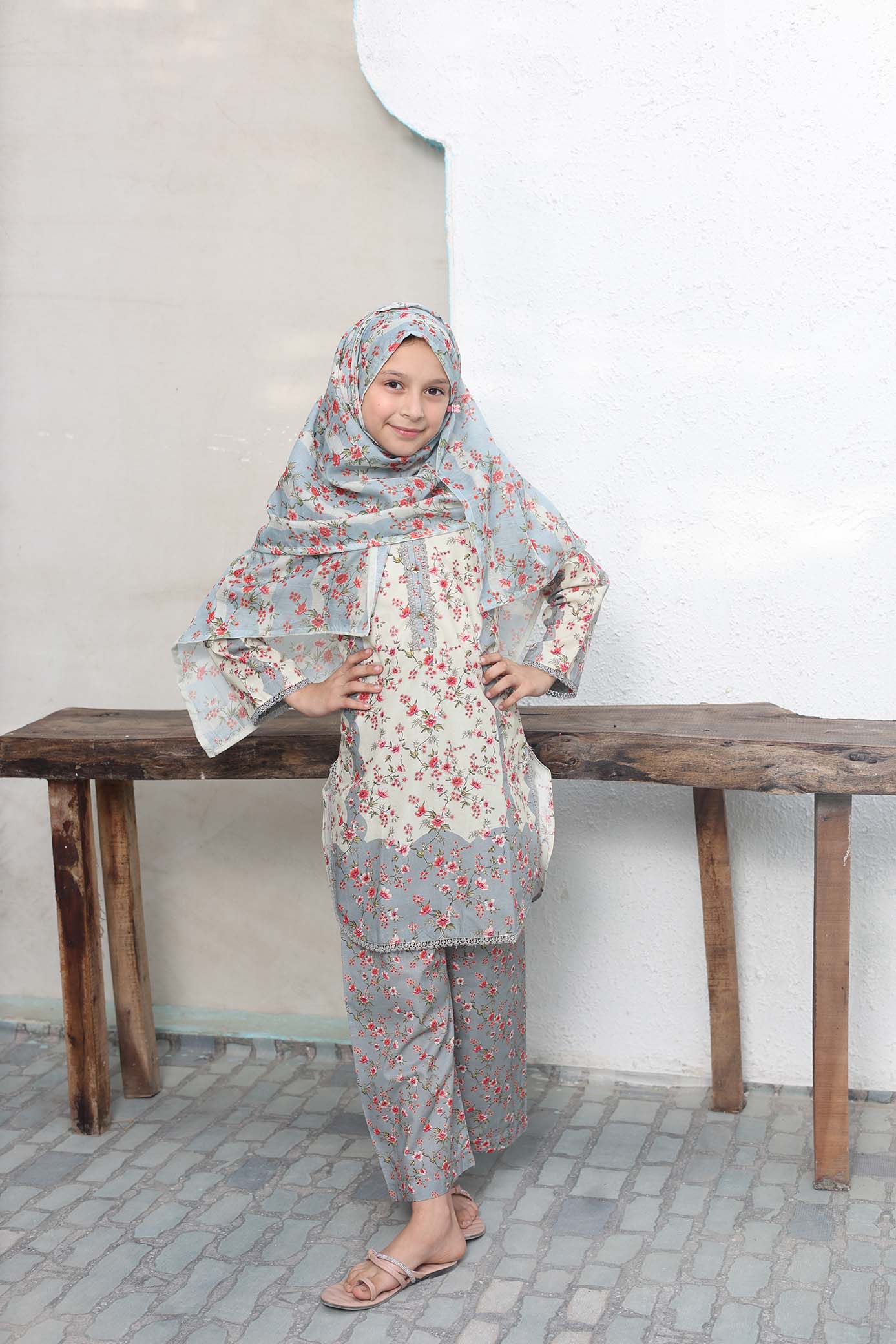 Young girl in a floral outfit standing on a wooden bench against a plain wall.