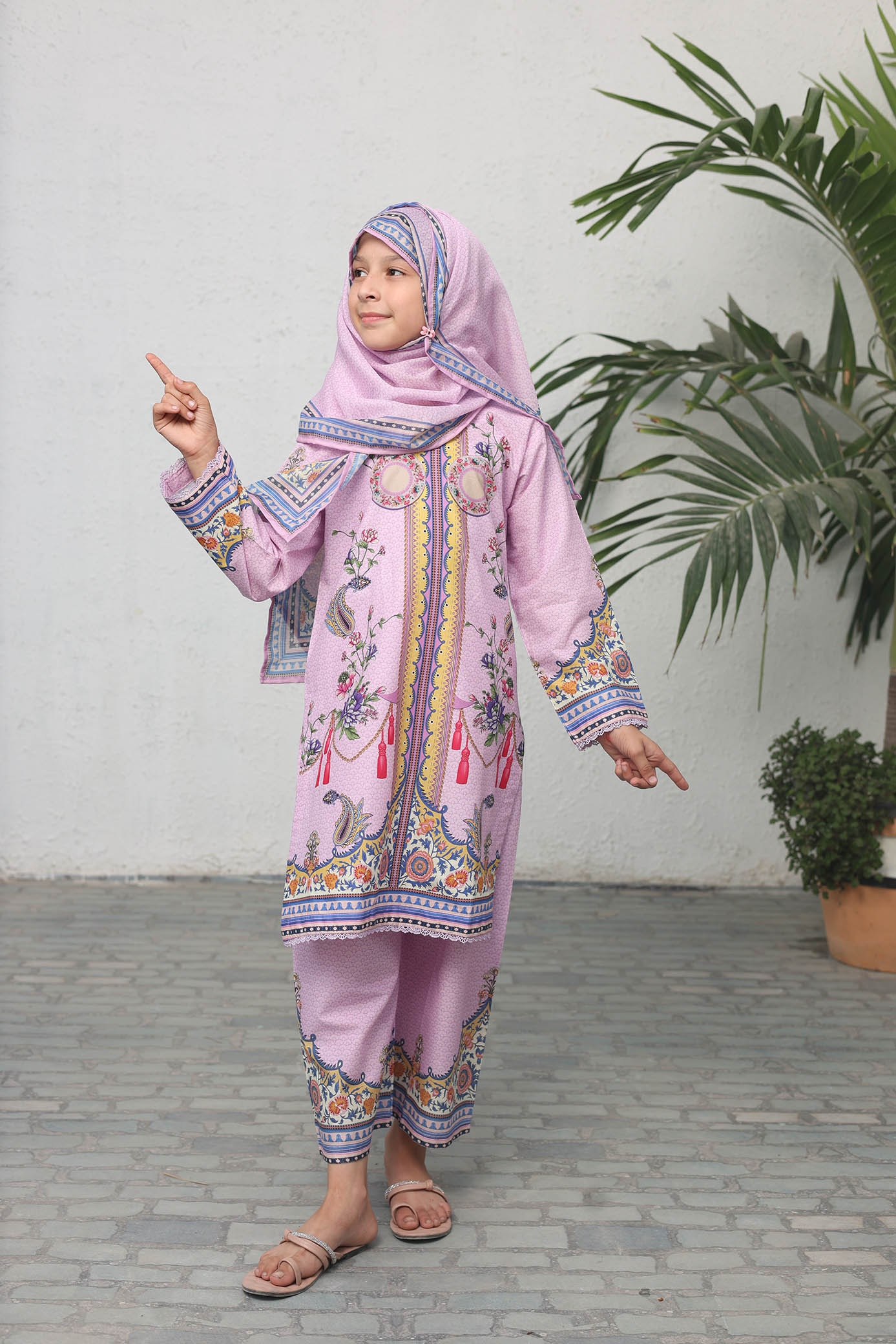Child wearing a pink traditional outfit with a headscarf, pointing towards the camera.