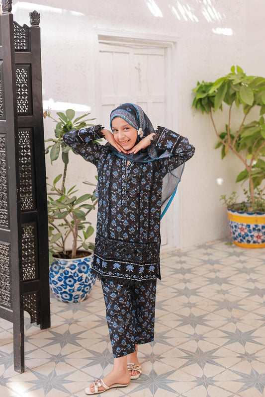 A woman posing in a black and blue embroidered festive outfit with long sleeves and a headscarf, standing in a room with decorative elements.