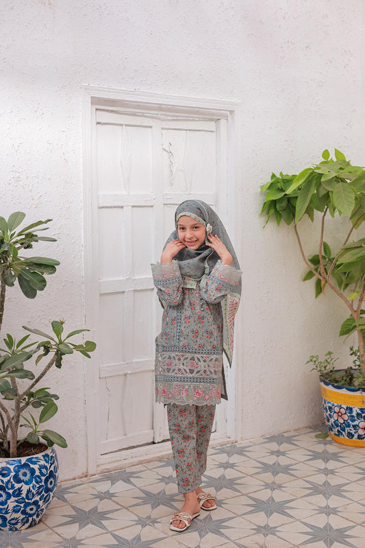 A woman standing in a room with a white door and decorative plants, wearing a floral and geometric patterned outfit with long sleeves and a dupatta.