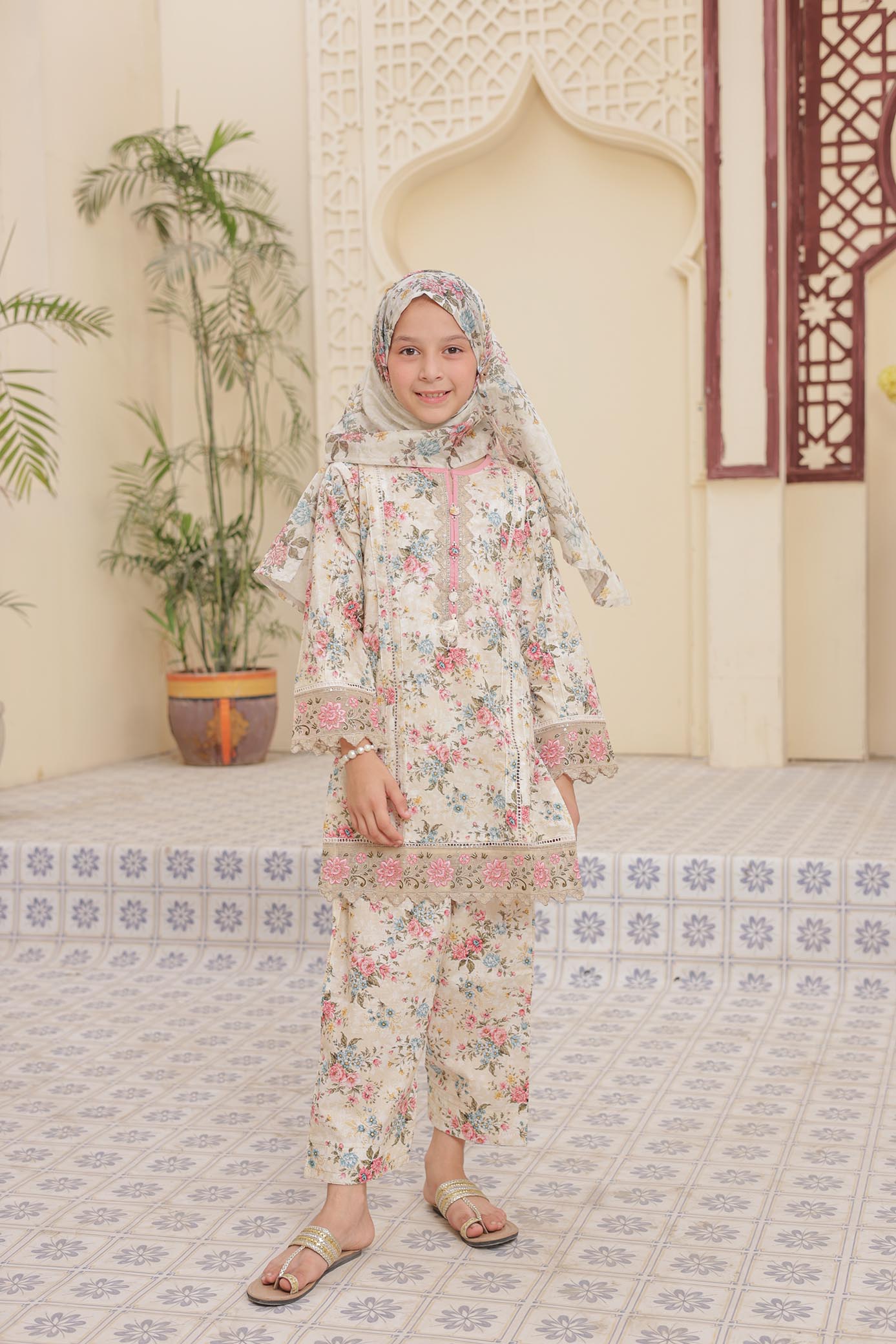 Young girl in a floral outfit standing in a decorated room with plants and architectural elements.