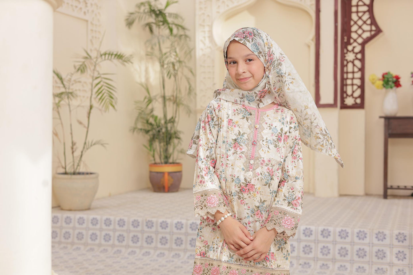 Young girl in a floral dress and headscarf standing in a decorated room with plants and furniture.