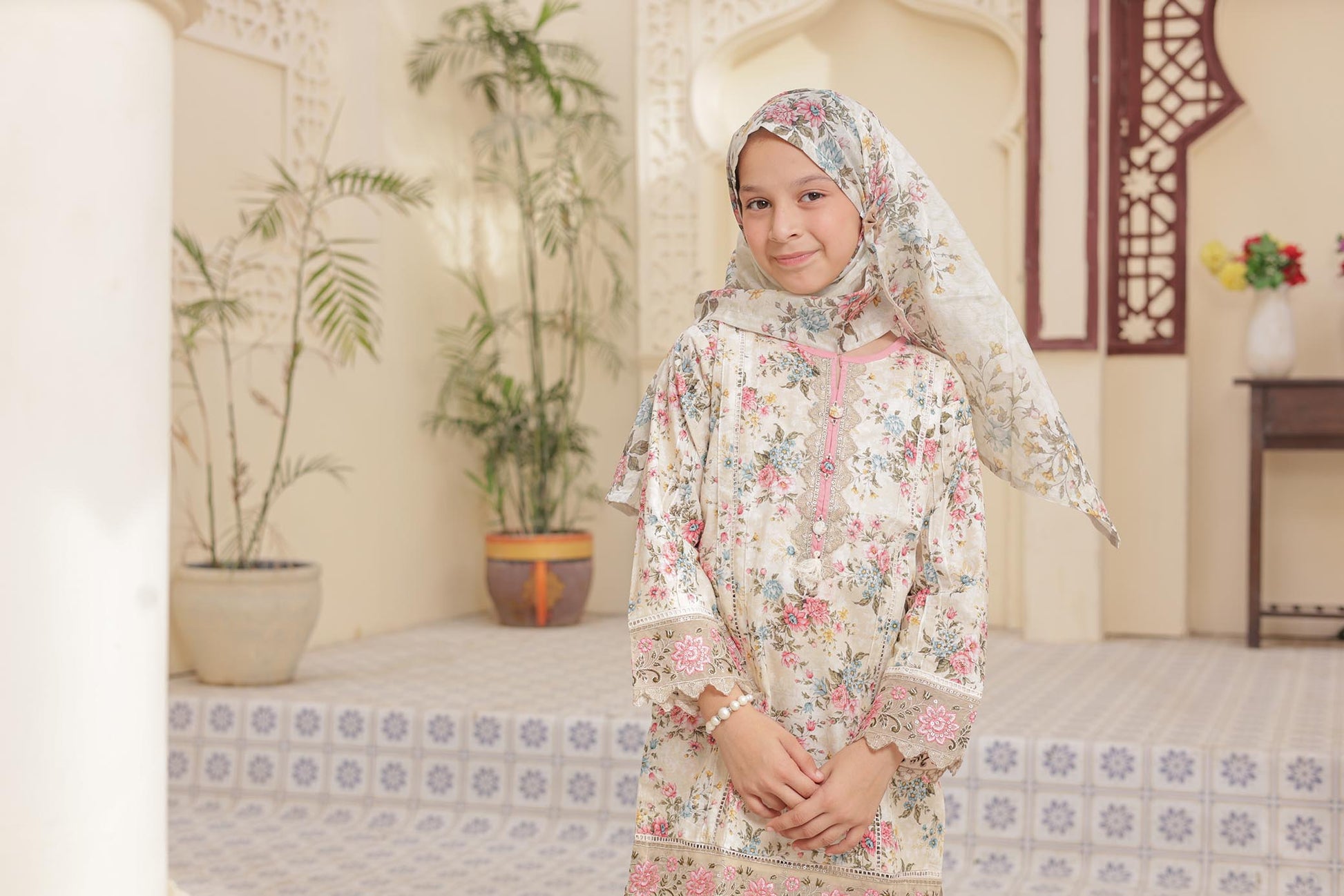 Young girl in a floral dress and headscarf standing in a decorated room with plants and furniture.