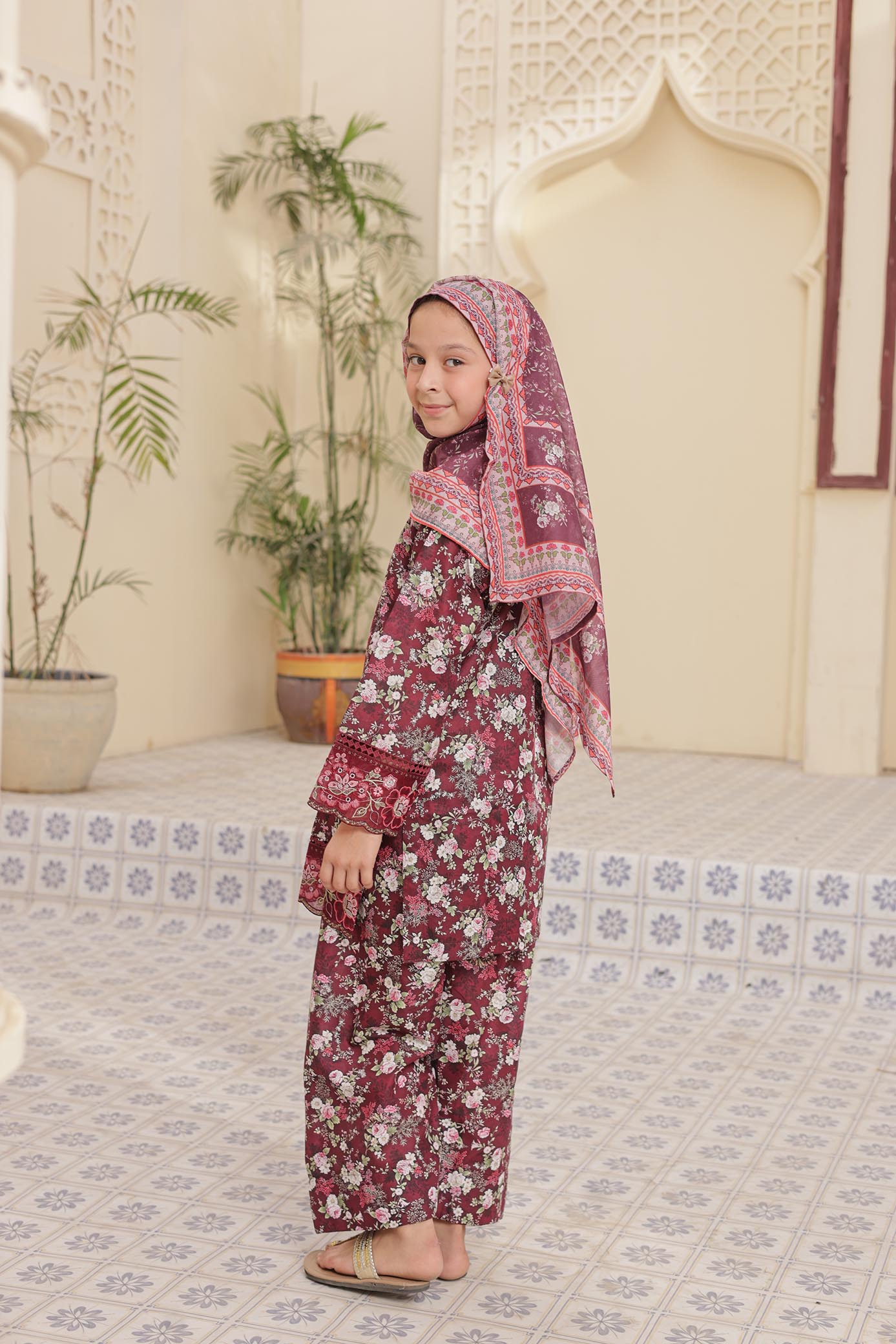 Young girl in a floral dress and headscarf standing in a decorated room with plants and a mirror.