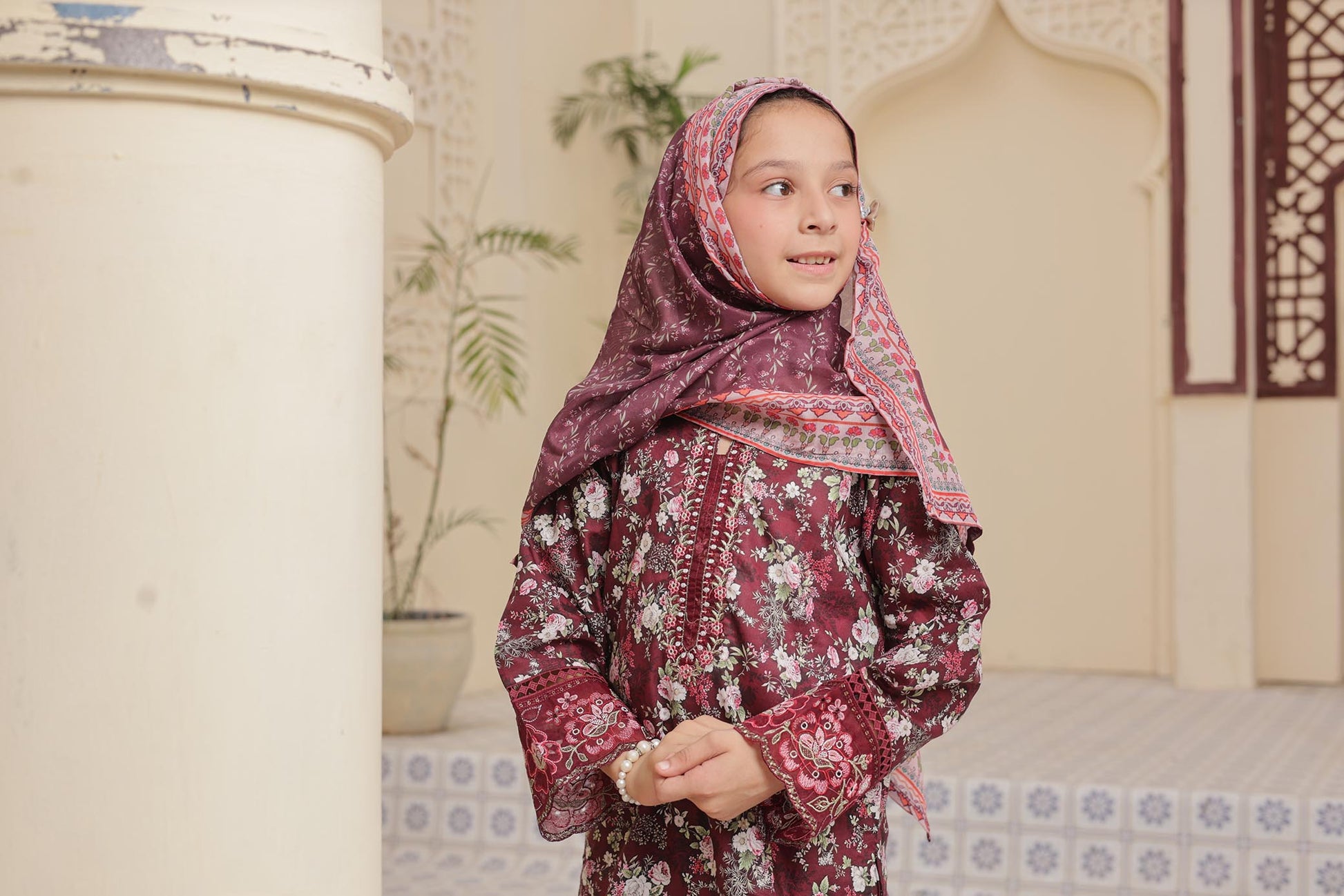 Young girl in a patterned dress and headscarf standing in a decorated room.