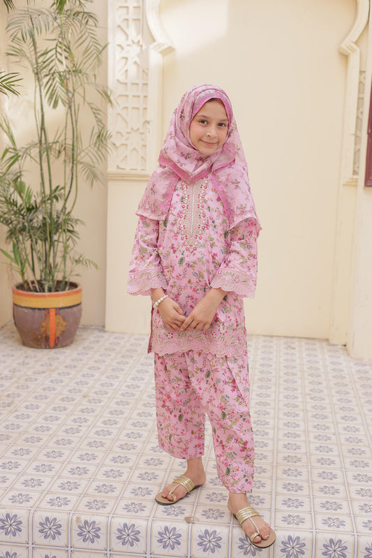 Young girl in a pink floral outfit standing on a patterned rug with a plant in the background.