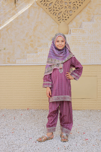 Young girl in a traditional outfit with a purple top and pants, standing against a beige wall.