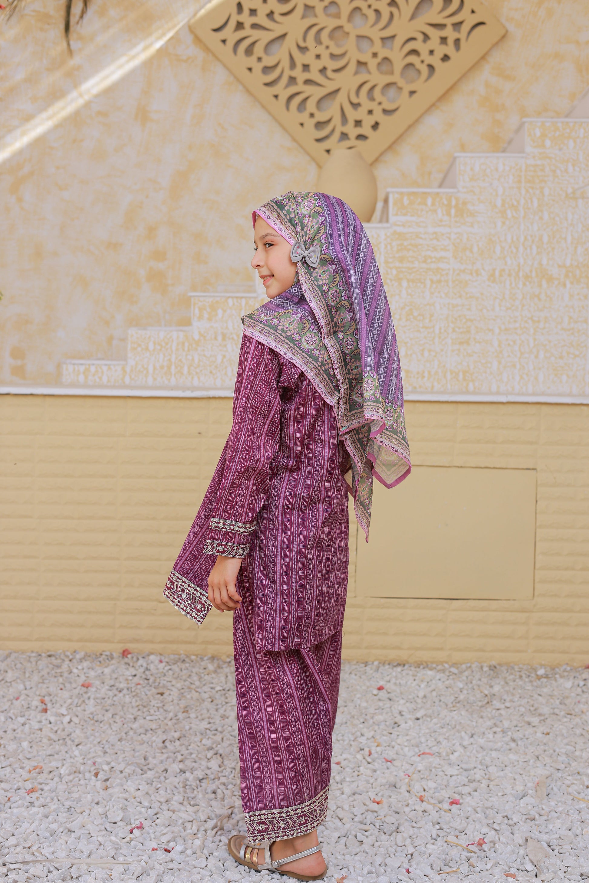 Woman in traditional purple outfit with patterned headscarf standing in front of a decorative wall.