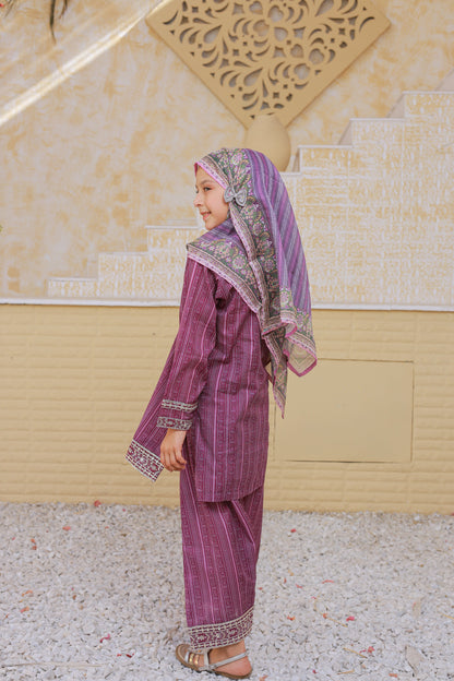 Woman in traditional purple outfit with patterned headscarf standing in front of a decorative wall.