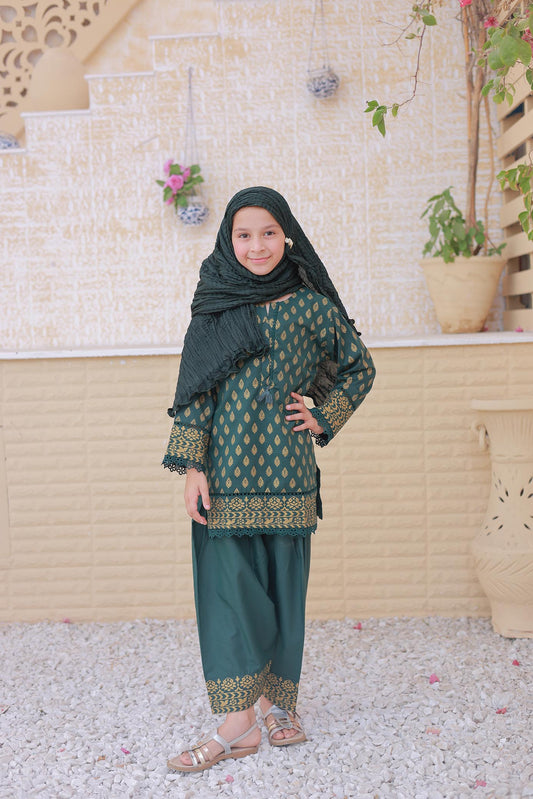Young girl in a green traditional outfit with a headscarf standing against a light-colored wall with plants.