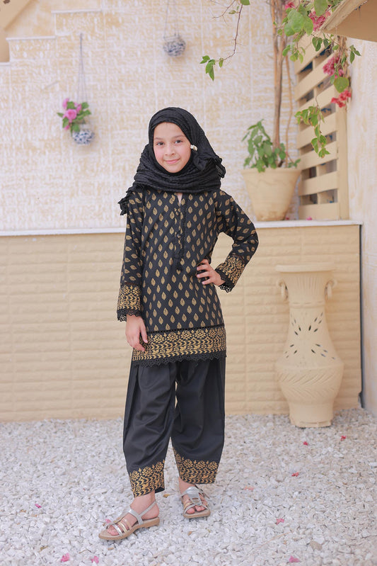 Young girl in a black and gold traditional outfit standing outdoors with decorative plants and a light-colored wall in the background.