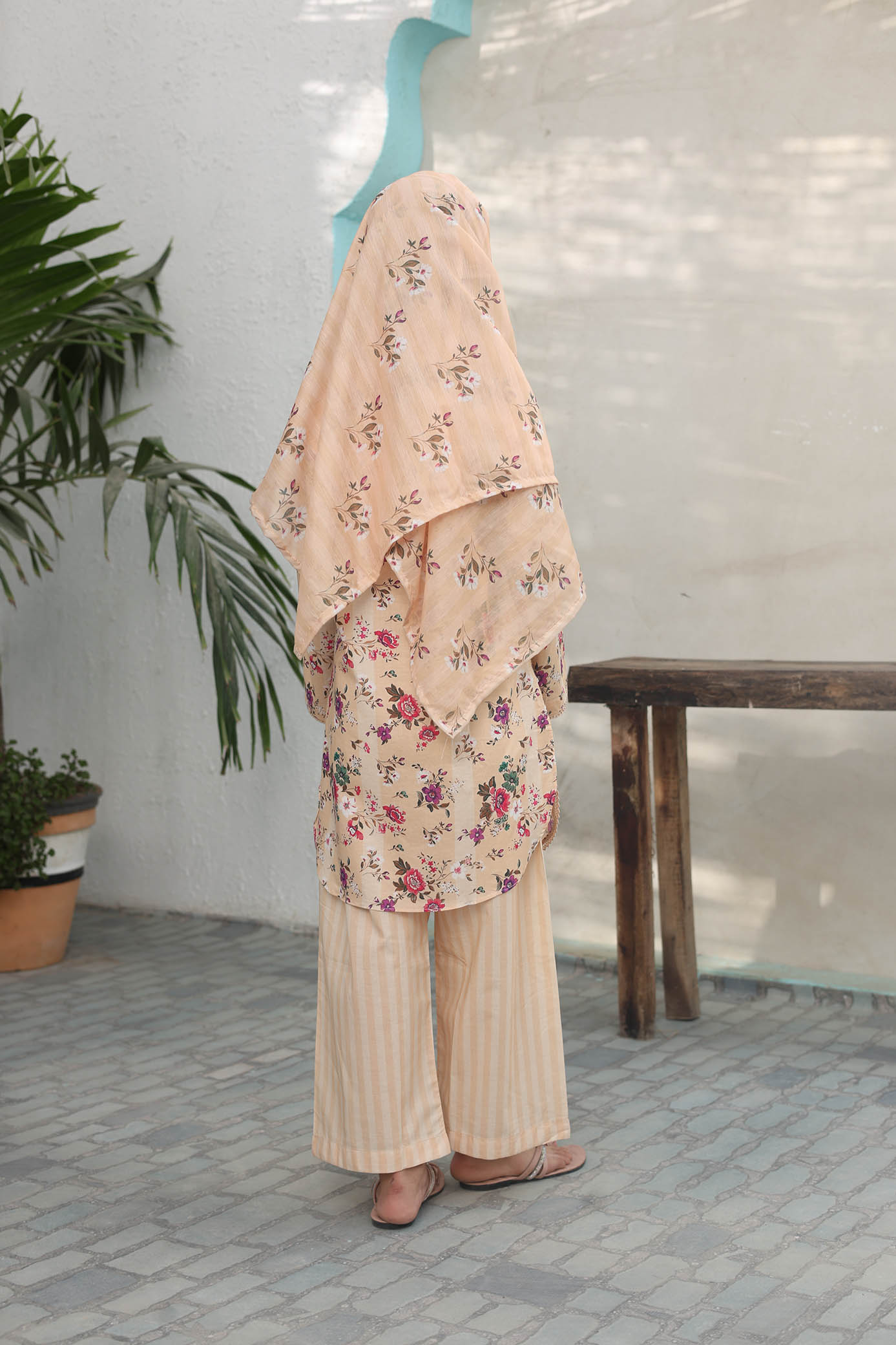 Woman wearing a floral dress and beige pants in an outdoor setting with a white wall and wooden table.