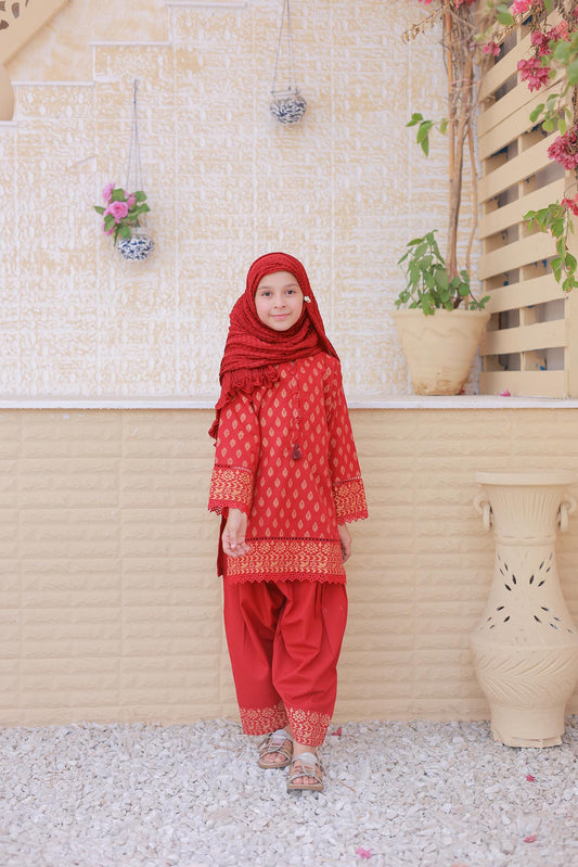 Child wearing a red traditional outfit standing in front of a beige wall with plants.