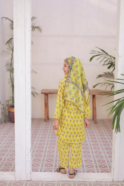 Woman in a yellow traditional outfit standing in a room with plants and a tiled floor.