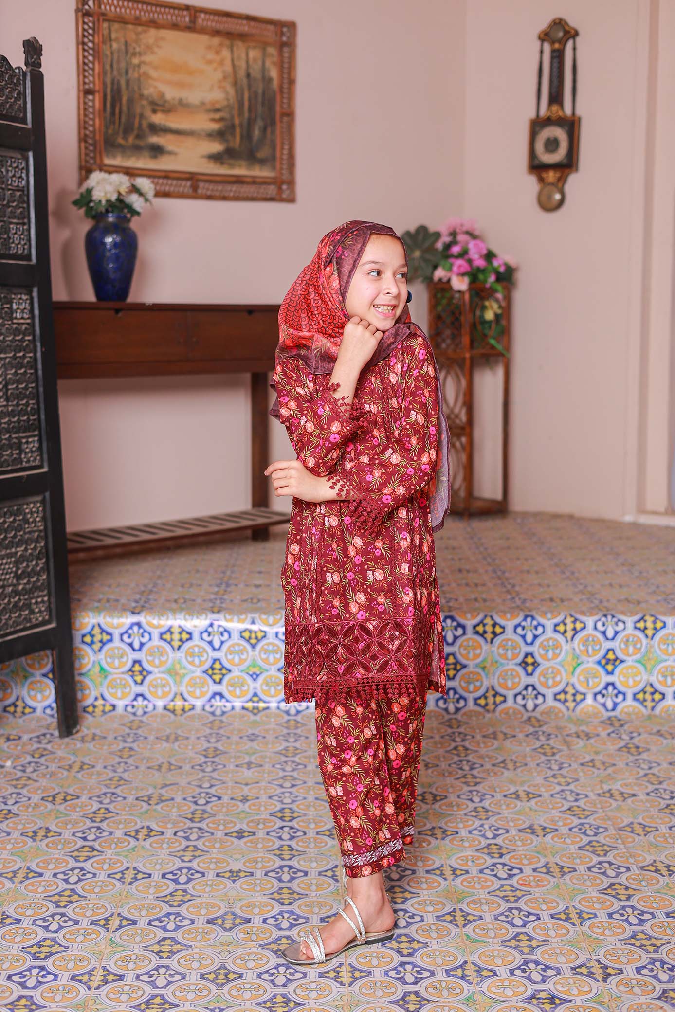 A girl wearing a red embroidered shirt with long sleeves, paired with a printed dupatta and matching trousers, standing in an indoor setting with decorative tiles on the floor.