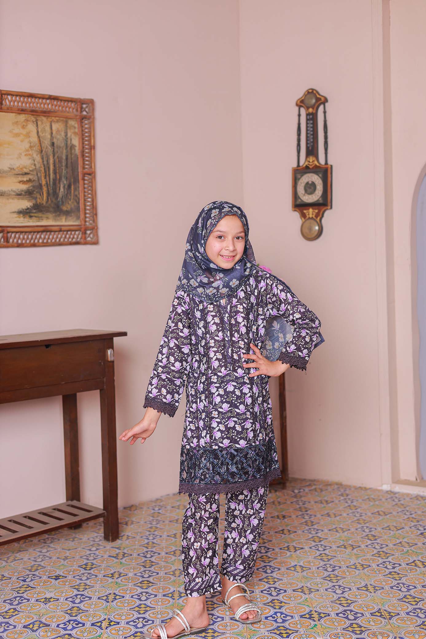 A girl wearing a long-sleeved embroidered shirt with laser lace cutwork, paired with a printed dupatta and trousers, standing in a room with a patterned floor.