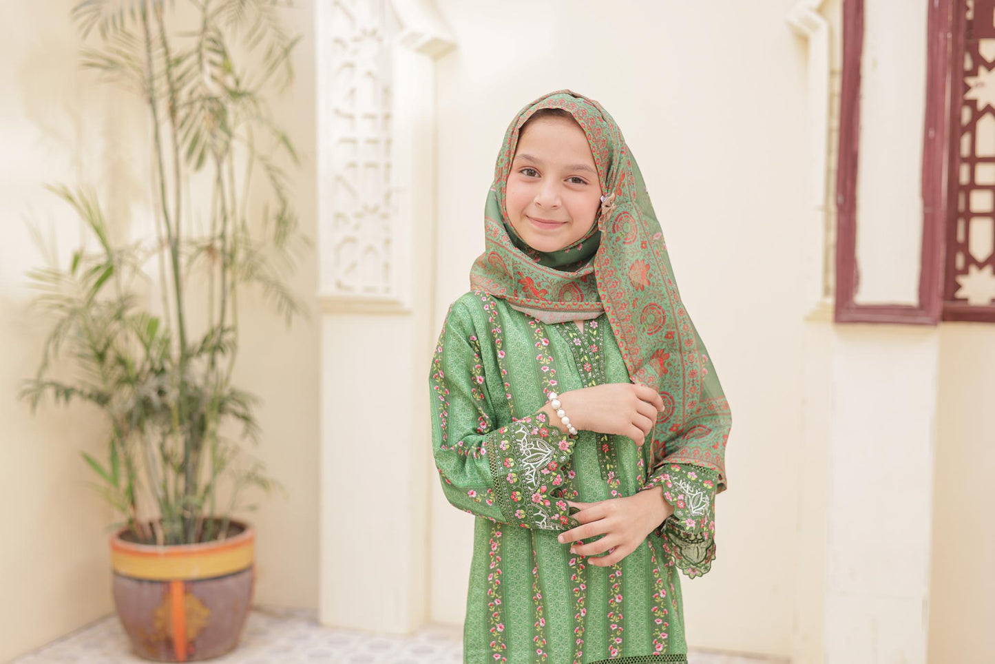 Young girl in a green traditional outfit with a headscarf standing indoors.