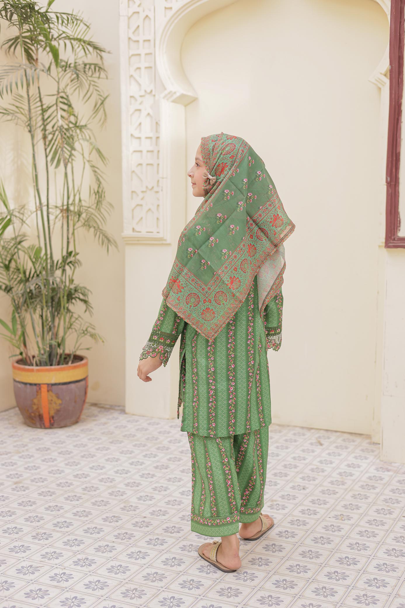 Woman in a green traditional outfit with a patterned shawl standing in a decorated room.