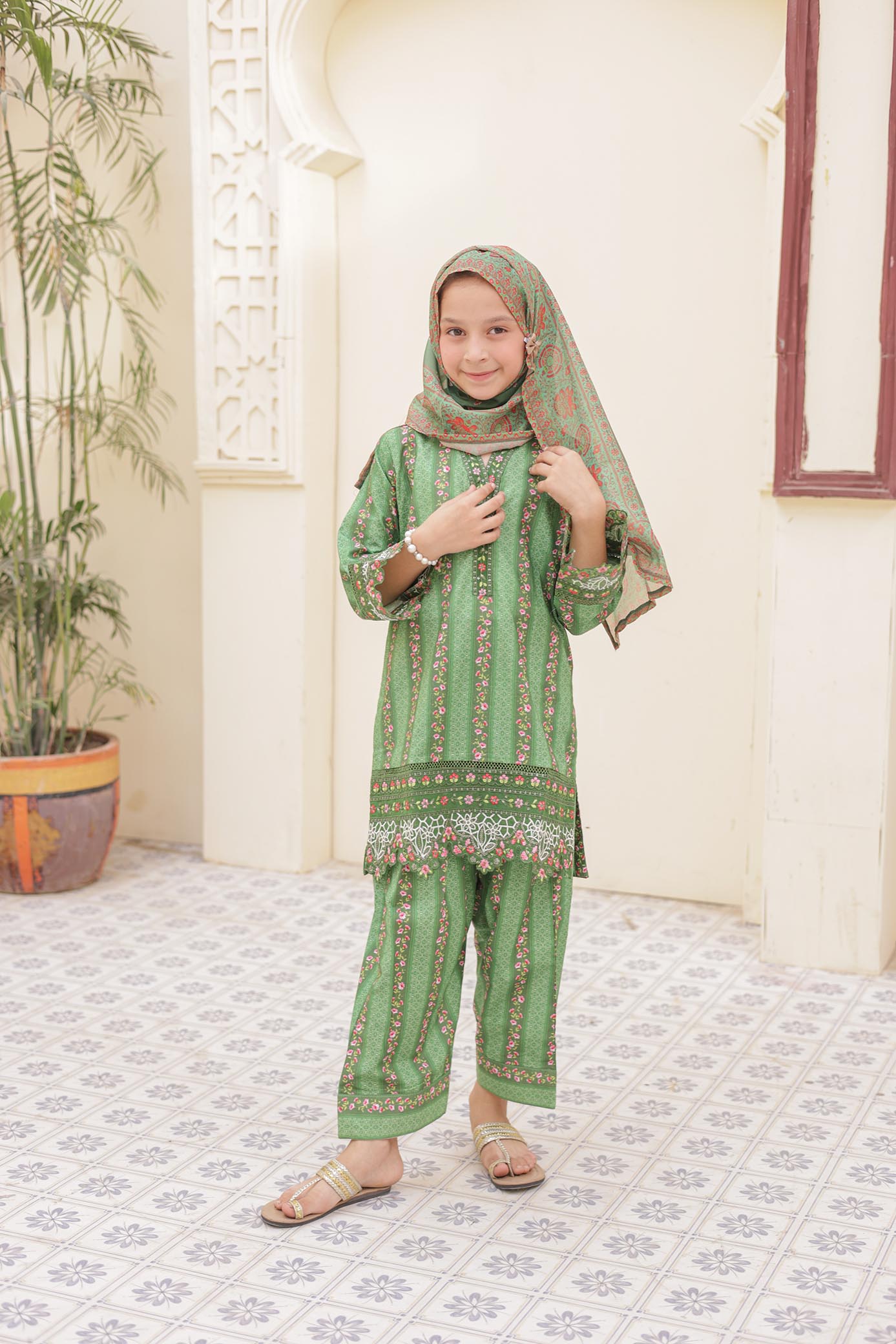 Girl in a green traditional outfit standing indoors with a plant and window in the background.