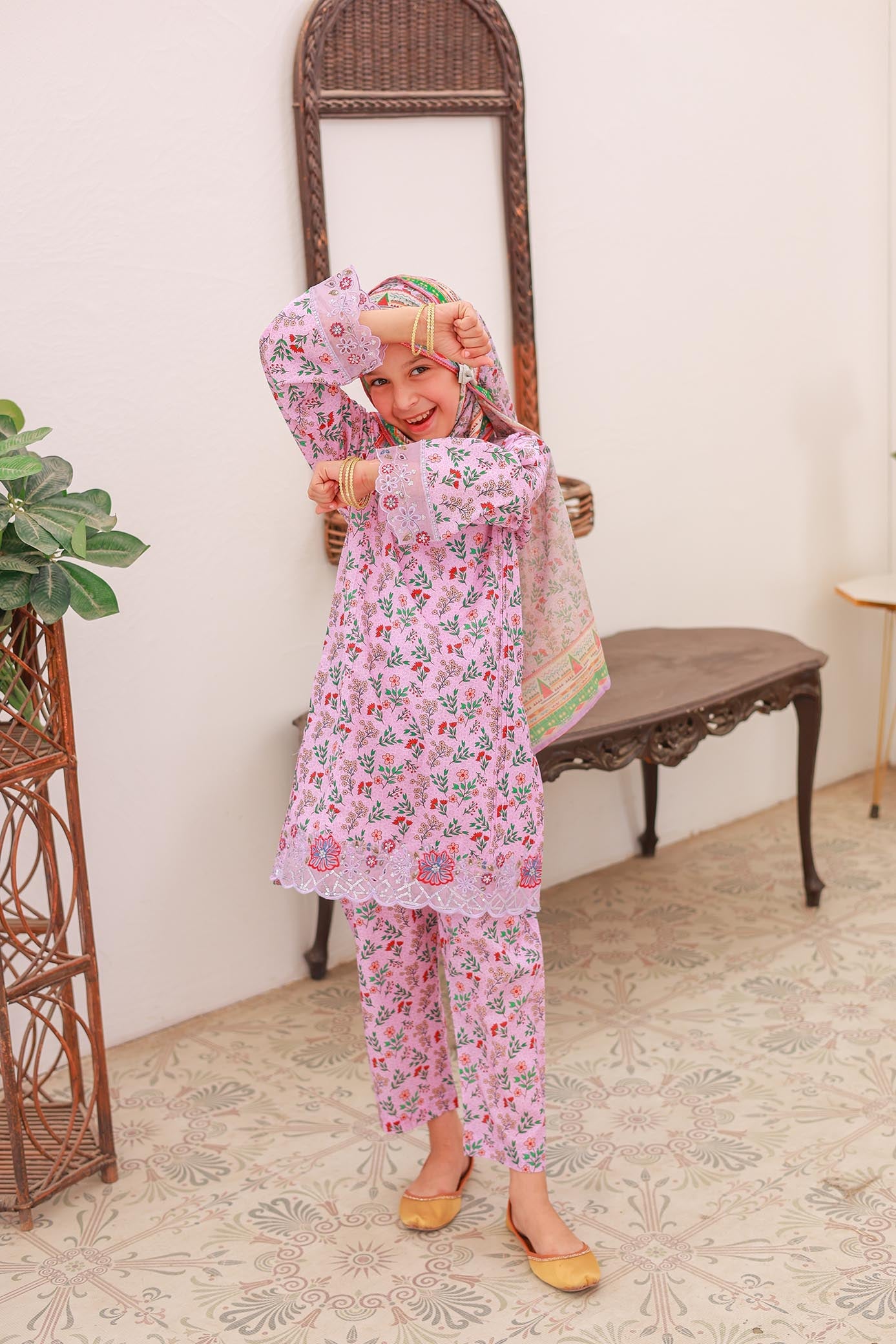 A girl wearing a festive pink three-piece outfit with embroidery and printed patterns, including a shirt, trousers, and a dupatta.