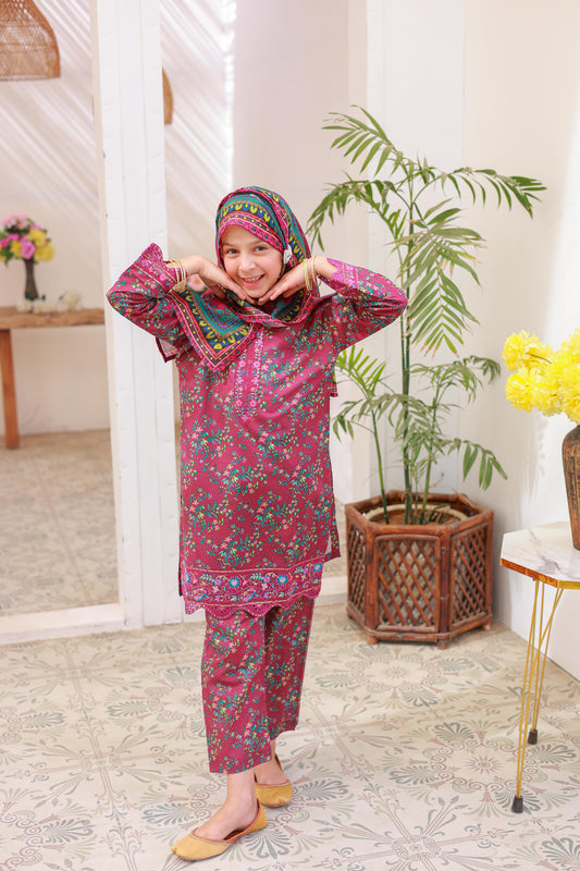 A girl wearing a traditional festive outfit with embroidery and lacework, consisting of a long-sleeved shirt, printed dupatta, and matching trousers, in a home setting with decorative plants in the background.